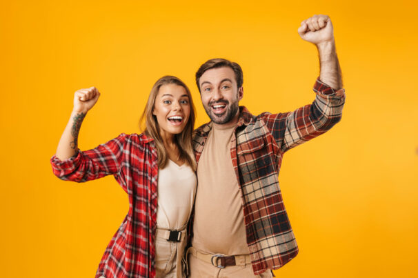 Beautiful excited couple hugging and making winner gesture isolated over yellow background